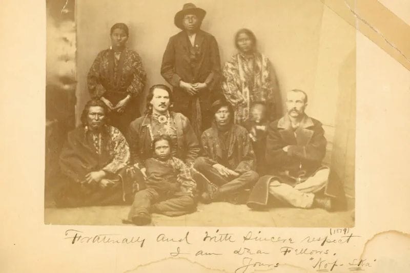 Sepia-toned historical group photograph showing Native American men, women, and children seated and standing together alongside a few non-Native individuals, dressed in period clothing, with handwritten notes visible beneath the image.