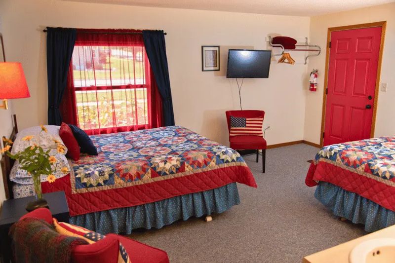 A guest room at A Guest Hus Motel features two beds with patriotic patchwork quilts, red-and-blue accents, a wall-mounted TV, and a bright red door, creating a cozy, Americana-inspired space.