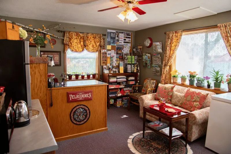 The welcoming office and lobby area at A Guest Hus Motel features cozy seating, travel brochures, local artwork, potted plants, and a small counter with a coffee station, creating a warm, homey atmosphere.
