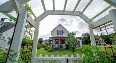 A Guest Haus Motel is viewed through a white garden arbor and picket gate, with the charming white-and-red house surrounded by greenery and a well-kept lawn on a bright day.