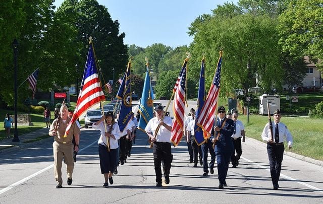 Members of the Lanesboro American Legion march down a sunlit street in a Memorial Day-style parade, carrying multiple American flags and service banners. The group includes veterans and uniformed participants walking in formation along the road, with green trees, parked cars, homes, and a few spectators in the background. The scene has a patriotic, community-focused feel, with bright spring or summer weather and small-town surroundings.