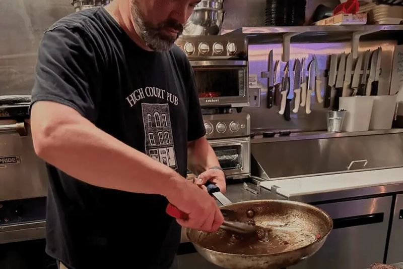 A cook prepares food in a restaurant kitchen, stirring a pan over the stove with professional equipment and knives visible, capturing behind-the-scenes cooking at a local pub.