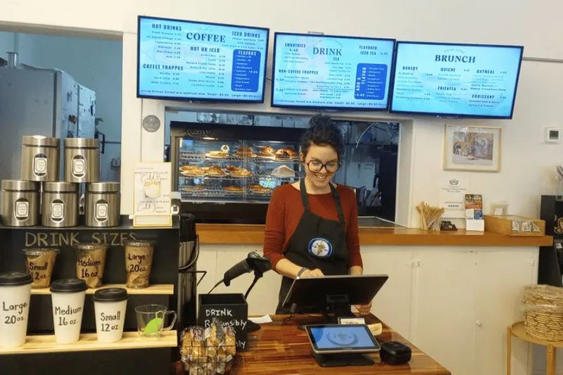 A barista smiles while taking an order at the Paddle On Coffee counter, with menu boards, pastries, and drink options displayed behind the register in a bright, welcoming café.