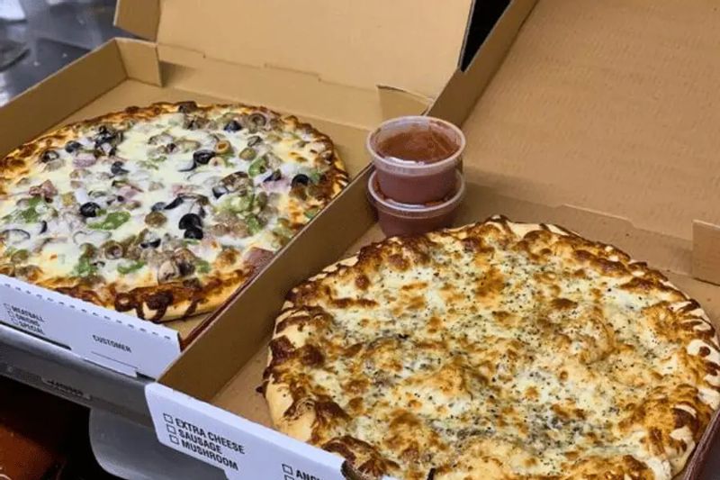 Two freshly baked pizzas sit in open takeout boxes, topped with melted cheese and assorted toppings, ready to enjoy from a local restaurant.
