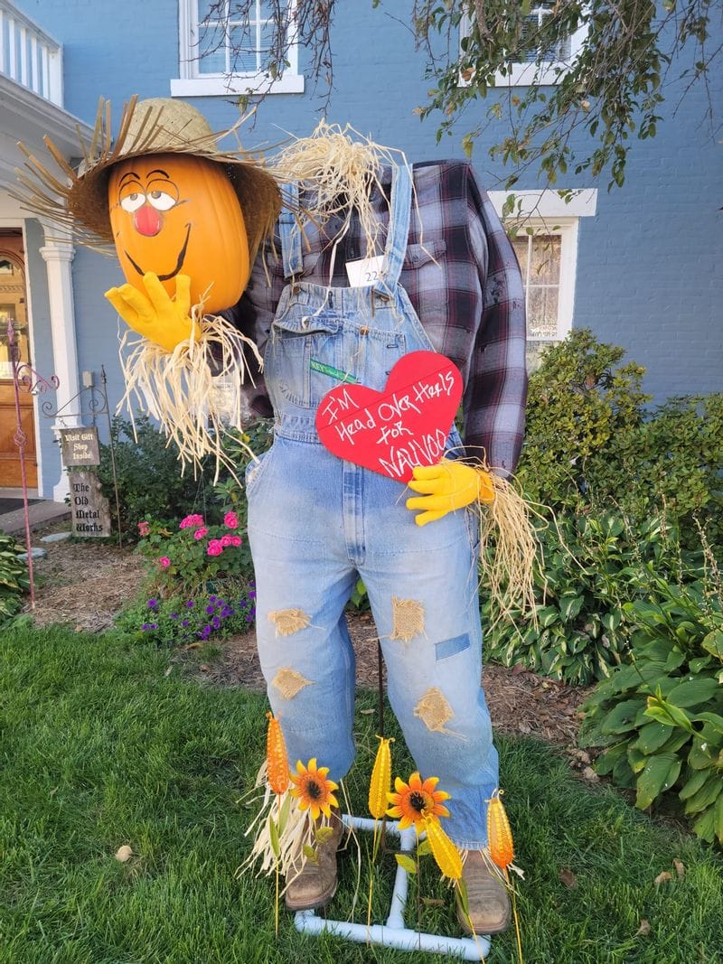 Headless scarecrow holding a smiling pumpkin head holding a heart that says "I'm head over heels for Nauvoo"
