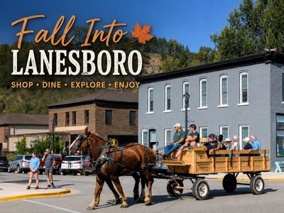 Promotional graphic for “Fall Into Lanesboro” showing a horse-drawn wagon ride traveling through downtown Lanesboro on a sunny day, with visitors seated in the wagon and the text “Shop • Dine • Explore • Enjoy” over a backdrop of bluffs and historic buildings.