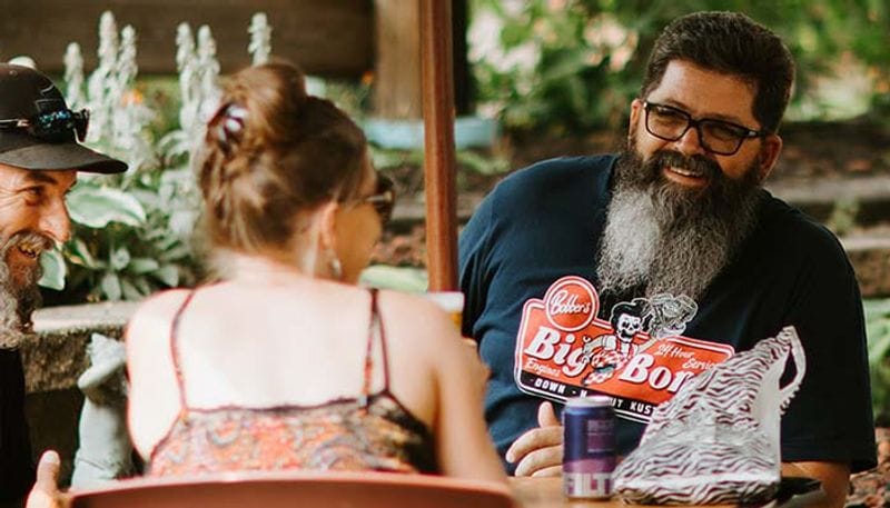 Friends sit together at an outdoor patio table in Lanesboro, smiling and chatting while enjoying drinks in a shaded garden setting.
