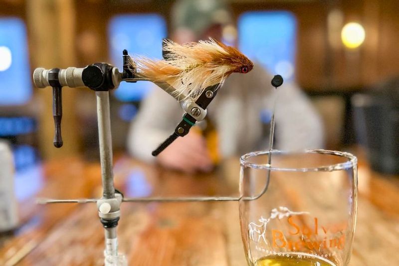 Close-up of a fly-tying setup at Sylvan Brewing in Lanesboro, with a tan-and-cream fishing fly clamped in a vise above a wooden table and a beer glass in the foreground, while a person sits blurred in the background.