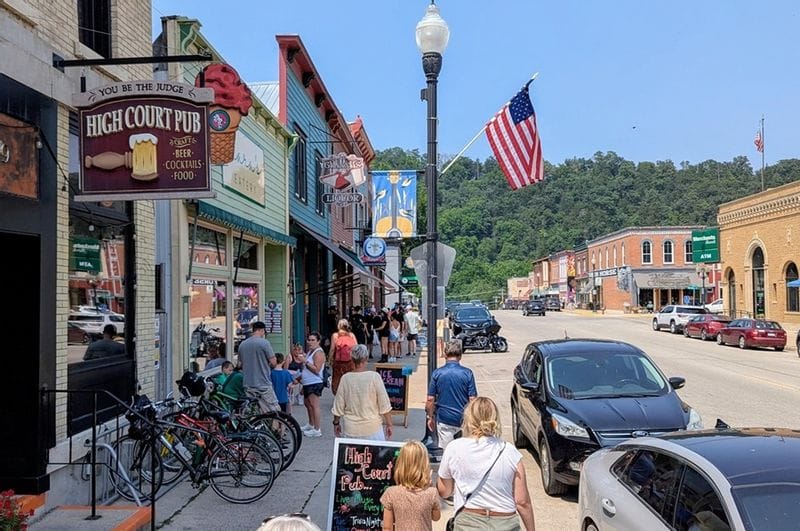 Visitors walk along Lanesboro’s bustling downtown sidewalk in summer, passing local shops, restaurants, bicycles, and parked cars with Bluff Country hills in the background.