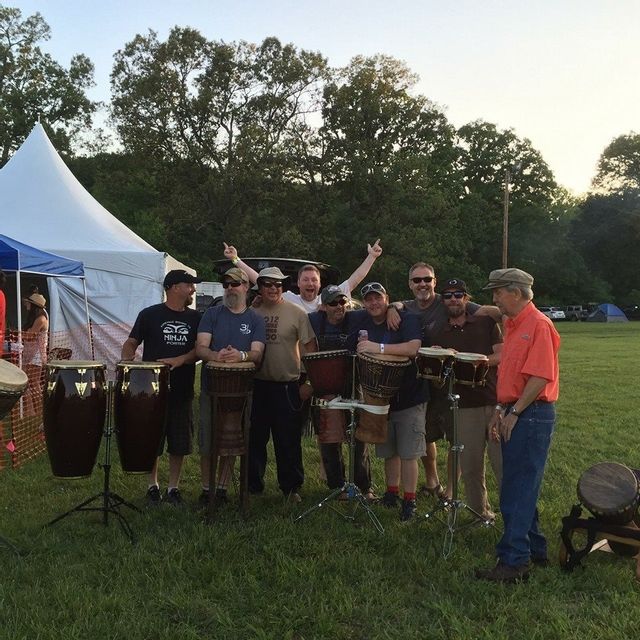 Members of the Iredell Rhythm Experience (IRE) stand together in a grassy outdoor area surrounded by hand drums and percussion instruments during Art in the Park. The group smiles and poses beside conga drums, djembes, and bongos near event tents as the sun begins to set behind the trees. The scene captures the fun and community spirit of the live music experience.