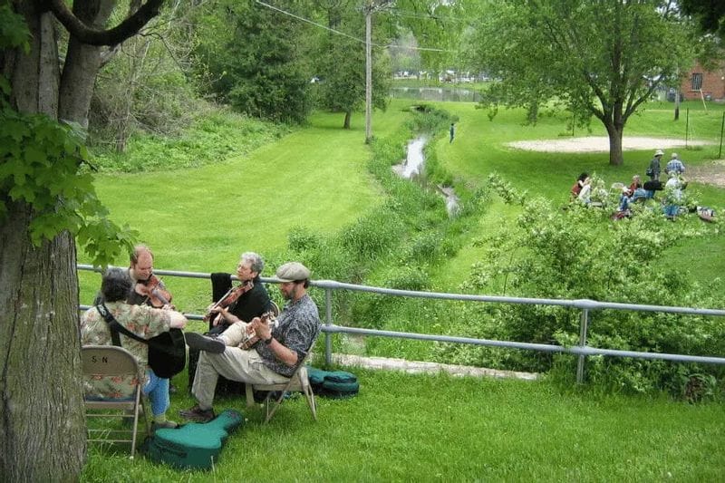 Musicians sit outdoors playing string instruments beside a grassy creek and park setting, with small groups of people gathered nearby during the Bluff Country Gathering in Lanesboro, MN