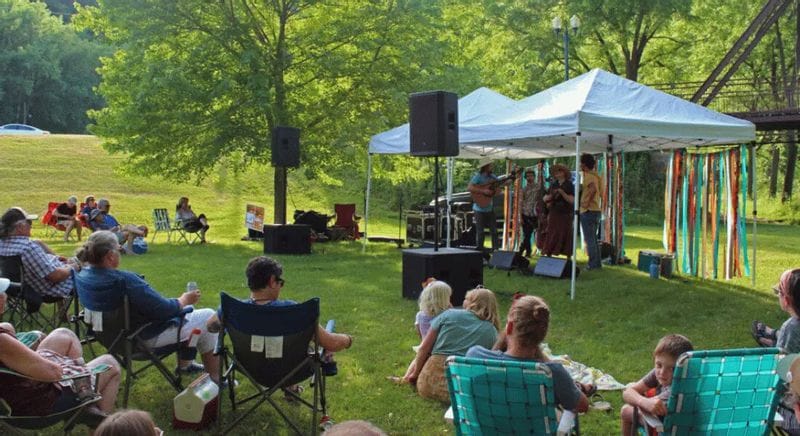 Audience seated on lawn chairs and blankets at an outdoor Rhythms on the Root concert in Lanesboro, Minnesota, watching a live band perform under a white canopy near the Root River with trees and a bridge in the background.