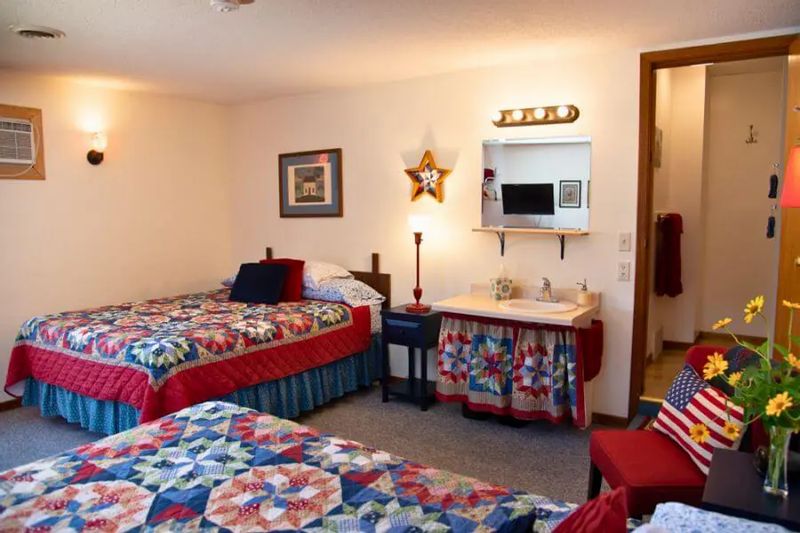 Bright guest room at A Guest Hus featuring two beds with colorful patchwork quilts, a small vanity sink area, patriotic décor accents, and an open doorway to the bathroom.