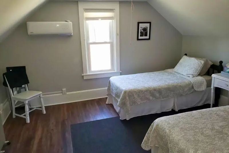 A simple bedroom inside a cozy cottage, featuring twin beds with light bedding, a sloped ceiling, wood floors, and a small window letting in natural light.