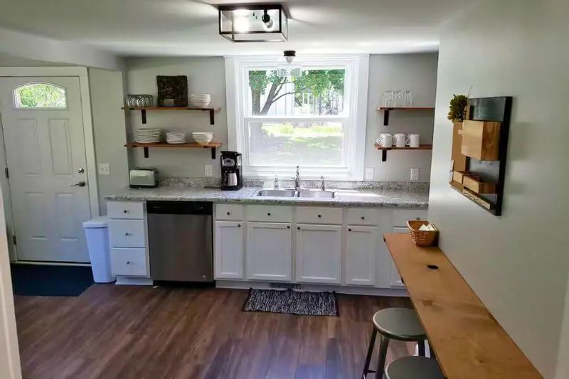 A bright kitchen inside a cozy cottage, featuring white cabinets, open shelving, a farmhouse sink beneath a window, and a small breakfast bar with stools.