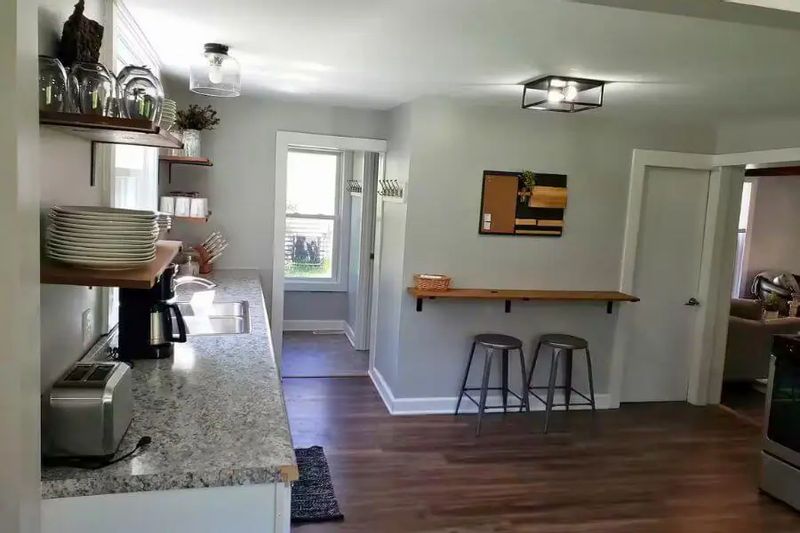 Another view of the cozy cottage kitchen, showing open shelving with dishes, a long countertop, and a small breakfast bar with stools.
