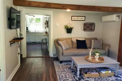A comfortable living room inside a cozy cottage, with a sofa, coffee table, wall-mounted TV, and an open doorway leading to the kitchen.