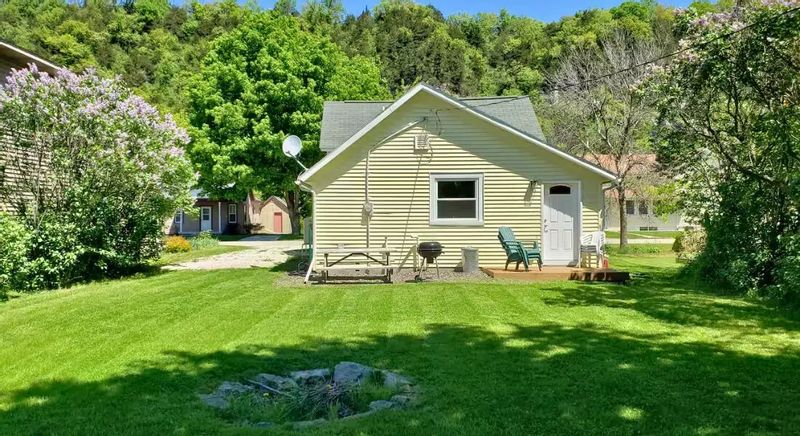 The backyard of a cozy cottage with a grassy lawn and fire pit, showing a small patio with chairs and a grill set against leafy hills on a sunny day.