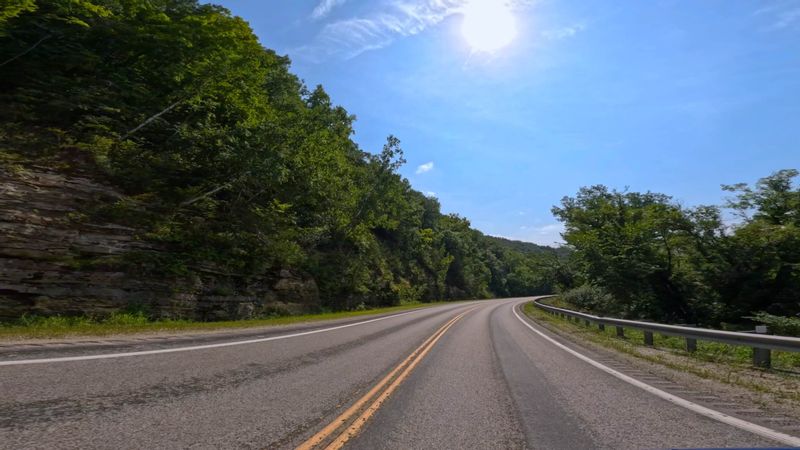 A winding stretch of the Historic Bluff Country National Scenic Byway curves through forested limestone bluffs under a bright blue sky on a sunny day in southeastern Minnesota.