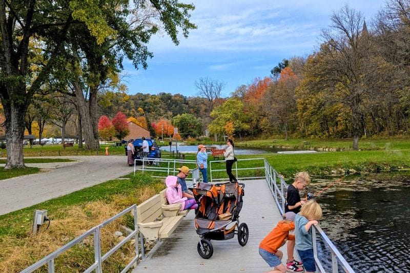Families fish from an accessible dock at Sylvan Park in Lanesboro, Minnesota, surrounded by colorful fall foliage along the Root River on a crisp autumn day.