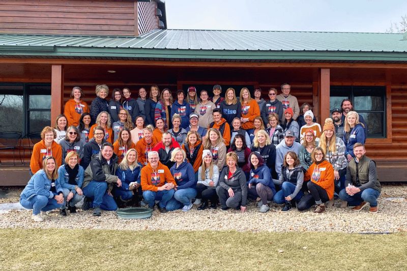 A large group of adults gathered outdoors in front of a log-style building at Cedar Valley Resort near Lanesboro and Whalan, Minnesota, posing together during a group retreat or conference on a cool-weather day.