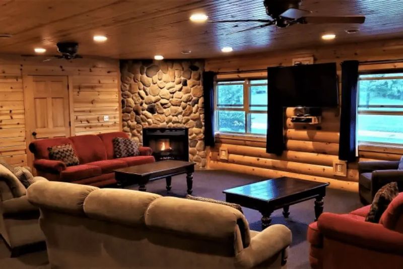 A cozy lodge-style living room at Cedar Valley Resort features log walls, a stone fireplace with a lit fire, comfortable sofas and armchairs, wooden coffee tables, and a mounted TV, creating a warm space for relaxing and gathering.