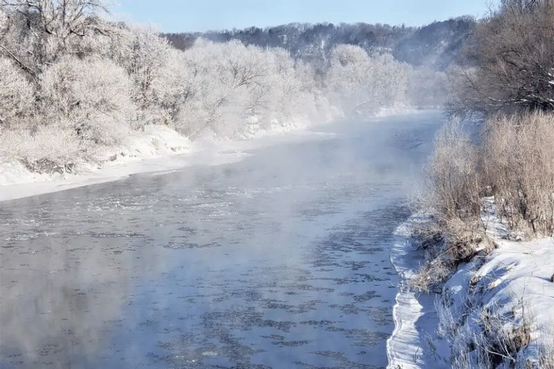 A winter view of the Root River in Whalan, Minnesota, near Cedar Valley Resort, showing a partially frozen river bordered by snow-covered banks and frost-coated trees, with light mist rising above the cold water.
