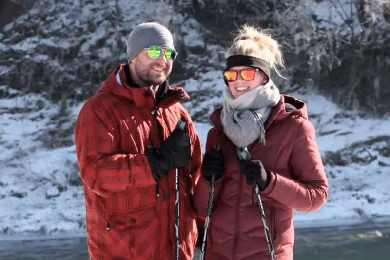 Two people dressed in winter jackets, hats, and gloves smile while standing with ski poles along a snowy trail near the Cedar Valley Retreat Center, enjoying a cold-weather outdoor activity with wooded bluffs in the background.