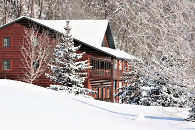 Cedar Valley Retreat Center is shown in winter as a red lodge-style building surrounded by snow-covered trees and rolling hills, with fresh snow blanketing the landscape in a peaceful wooded setting.