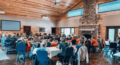 A large group gathers for a meeting or retreat inside the Cedar Valley Retreat Center, seated at tables in a spacious lodge-style room with wood-paneled walls, a stone fireplace, and large screens at the front.