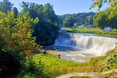 Lanesboro Dam spilling into the Root River on a sunny fall day, surrounded by trees and grassy riverbanks with a few people standing near the water below the dam.