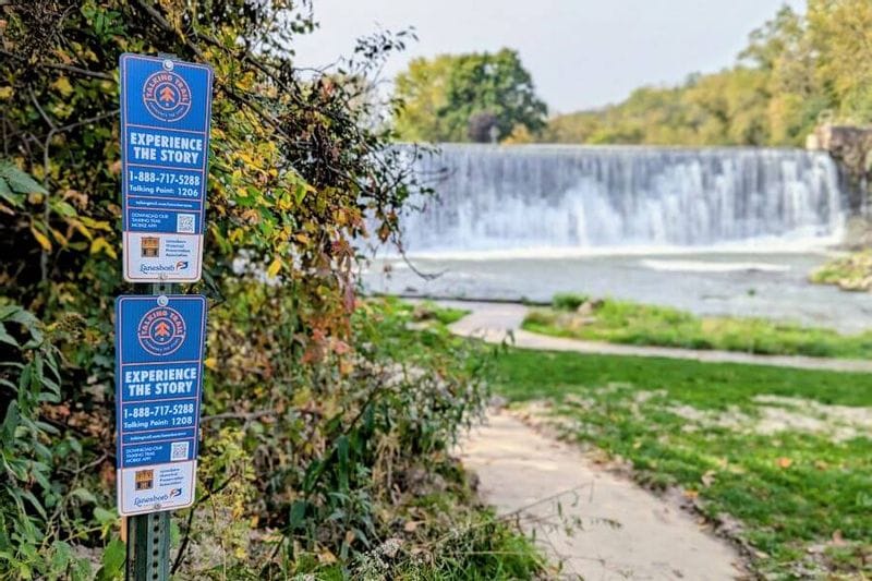 Lanesboro Talking Trail signs stand beside a leafy path near the Lanesboro dam, with water cascading over the spillway in the background along the Root River.