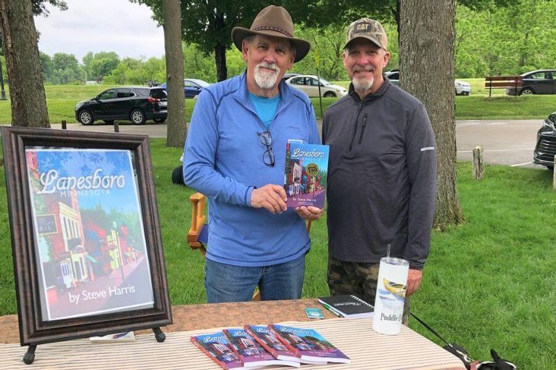 Author Steve Harris poses with another man at an outdoor event in Lanesboro, Minnesota, holding his book Lanesboro, with copies displayed on a table and a framed book cover beside them.