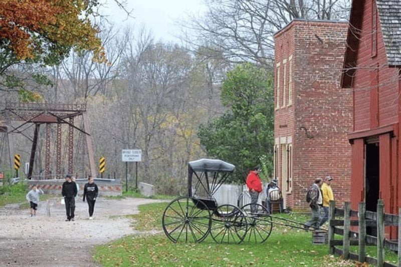 Visitors walk through Forestville State Park past historic brick buildings and a horse-drawn wagon, with a metal truss bridge and wooded landscape visible in the background during autumn.