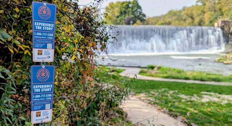 Lanesboro Talking Trail signs along a riverside path near the Lanesboro dam, with the Root River flowing past a wide waterfall and early fall foliage in the background.