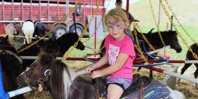 A child with face paint rides a pony on a small carousel during Buffalo Bill Days, with other ponies and the tent structure visible in the background.
