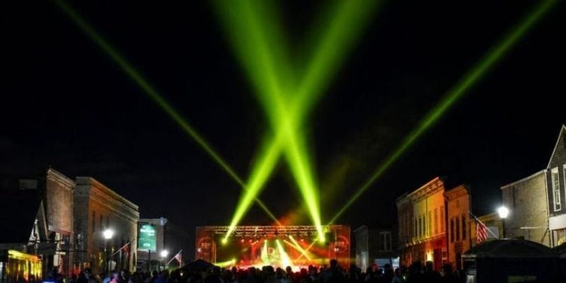 A nighttime Buffalo Bill Days street concert in downtown Lanesboro, Minnesota, with a large crowd gathered between historic storefronts. Bright green and yellow stage lights sweep high into the dark sky above the outdoor stage, while American flags, streetlamps, and illuminated buildings frame the lively festival scene.