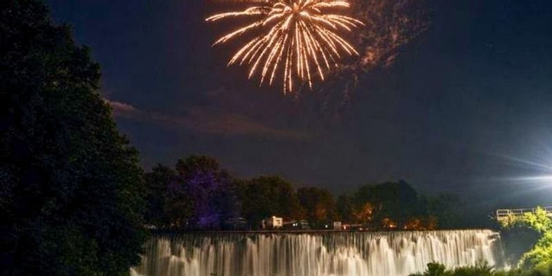 A large golden firework bursts over the illuminated Lanesboro Dam at night, with water cascading across the dam below and dark trees framing the scene.