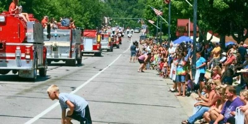 Children and families line the street during a summer parade in Lanesboro as fire trucks drive past, with a young boy running into the road to pick up candy while spectators watch from the curb.