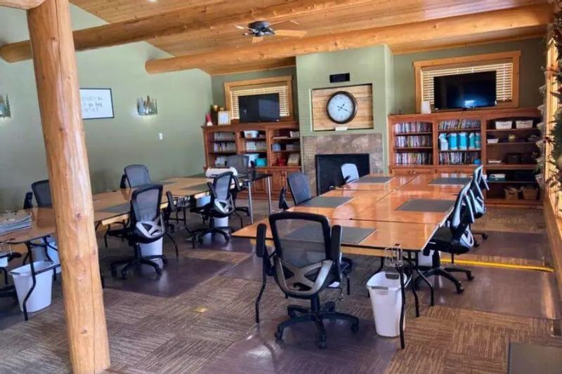 A spacious meeting room with long wooden conference tables, rolling office chairs, exposed wood beams, a stone fireplace with a clock, bookshelves, and wall-mounted TVs, set up for a retreat or group meeting.