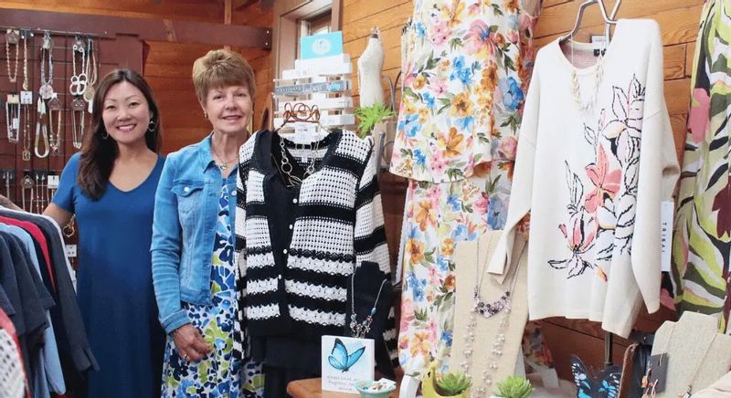 Two women stand inside a Lanesboro boutique surrounded by colorful dresses, sweaters, jewelry, and accessories, highlighting local shopping and small-town style.