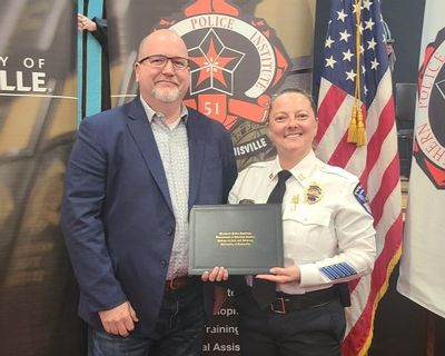 Captain Samantha Lindon of the Statesville Police Department stands beside SPD Police Chief Onley while holding her graduation certificate from the Southern Police Institute leadership program. Captain Lindon is dressed in her police uniform, and Chief Onley wears a blue blazer and glasses. The pair smiles for the photo in front of police institute banners and an American flag.