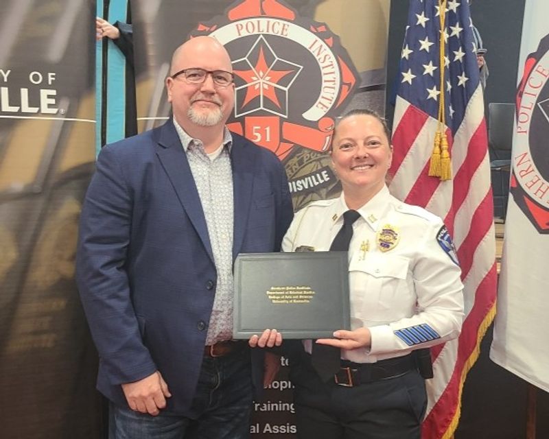Captain Samantha Lindon of the Statesville Police Department stands beside SPD Police Chief Onley while holding her graduation certificate from the Southern Police Institute leadership program. Captain Lindon is dressed in her police uniform, and Chief Onley wears a blue blazer and glasses. The pair smiles for the photo in front of police institute banners and an American flag.