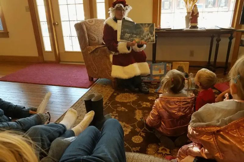 Mrs. Claus sits in an upholstered chair reading a Christmas storybook aloud to a small group of children seated on the floor, gathered closely around her inside a cozy, warmly lit historic home decorated for the holidays.