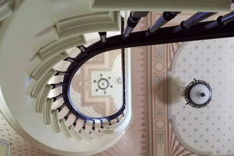 Looking up a curved staircase inside the Thompson House reveals ornate Victorian ceilings, decorative plasterwork, and historic light fixtures in a beautifully preserved interior.
