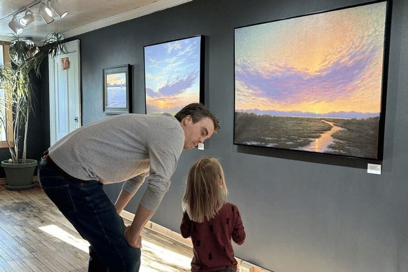 An adult and child stand together inside a Lanesboro art gallery, looking closely at colorful landscape paintings displayed on the wall under gallery lighting.