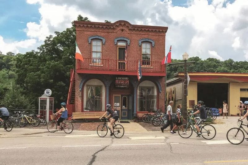 Cyclists ride past the Lanesboro History Museum, a historic brick building downtown, with bicycles parked outside and visitors walking along the sidewalk on a summer day.