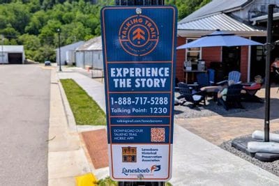 Talking Trail sign in downtown Lanesboro reading “Experience the Story,” with phone number and QR code, placed along a sidewalk near outdoor seating and local businesses.
