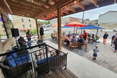 Live music performance on a covered patio at Sylvan Brewing, with musicians playing while people gather at outdoor tables under colorful umbrellas in downtown Lanesboro.