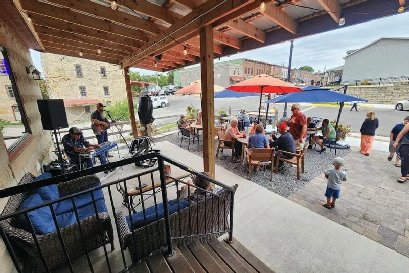 Live music performance on a covered patio at Sylvan Brewing, with musicians playing while people gather at outdoor tables under colorful umbrellas in downtown Lanesboro.